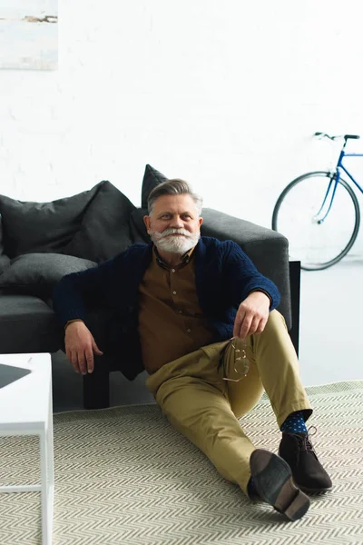 Knappe Glimlachend Senior Man Bedrijf Brillen Zittend Tapijt Thuis — Stockfoto
