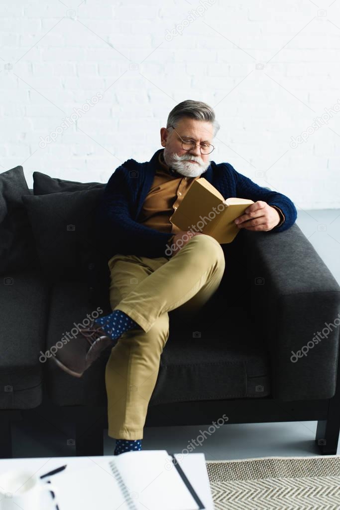 stylish bearded man reading book on sofa at home