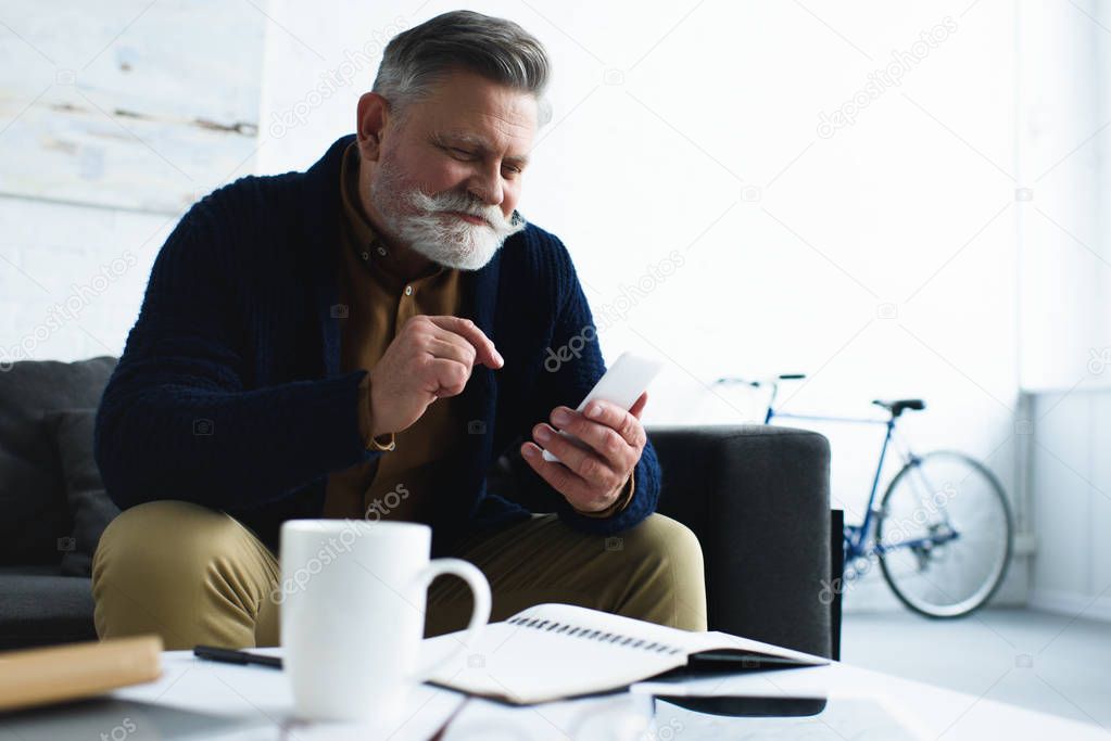 handsome smiling senior man using smartphone while sitting on sofa at home 