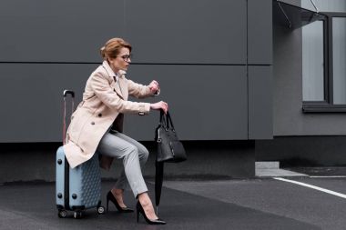businesswoman in stylish coat sitting on suitcase while waiting for taxi on street clipart