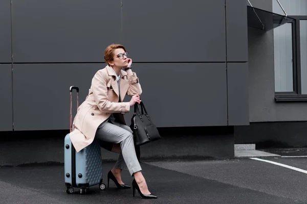 穿着时髦大衣的女商人坐在手提箱里等待出租车上街 — 图库照片