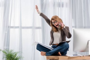 happy businesswoman with papers talking on smartphone while sitting on table in office clipart