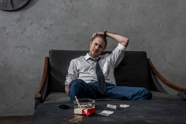 depressed businessman with drug addiction sitting on couch in front of table with drugs