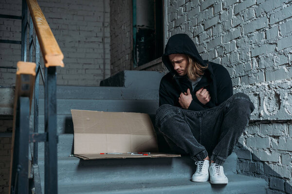 hooded heroin addicted junkie sitting on stairs with syringes on cardboard