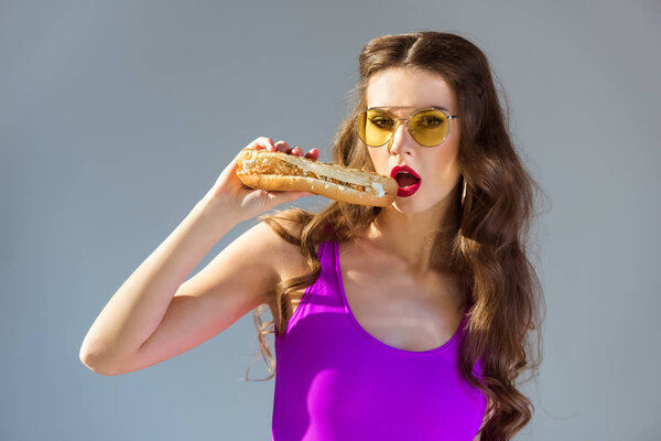 sexy attractive girl in ultra violet swimsuit eating hot dog and looking at camera isolated on grey