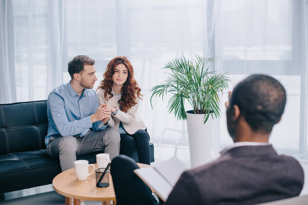 Young couple sitting on sofa in office and psychiatrist with textbook