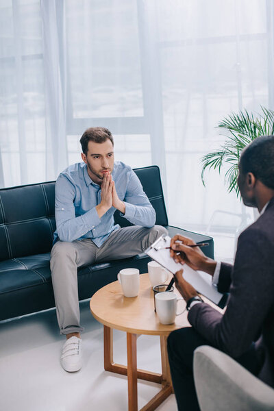 Young man sitting on couch and talking to african american counselor
