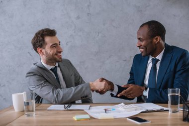 Multicultural business partners shaking hands at working table with documents  clipart