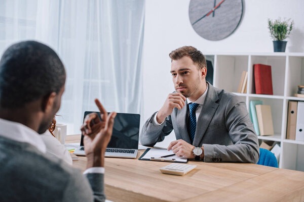 Агент по недвижимости сидит за столом с ноутбуком и слушает клиента
