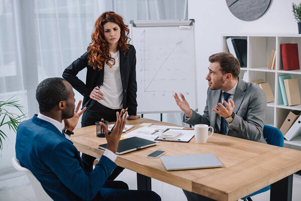 Financial adviser standing near table with laptop and papers while two businessmen arguing with her