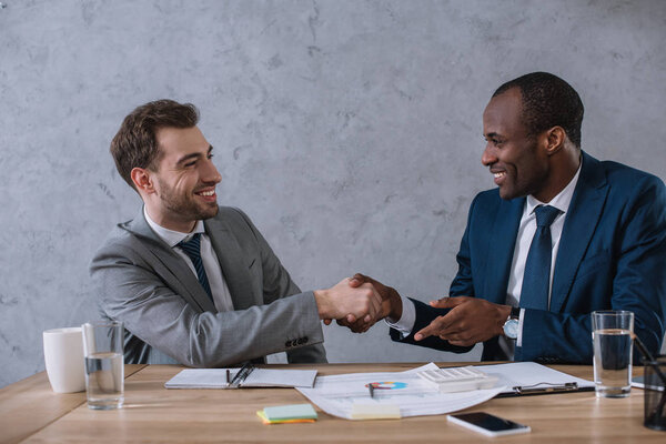 Multicultural business partners shaking hands at working table with documents 