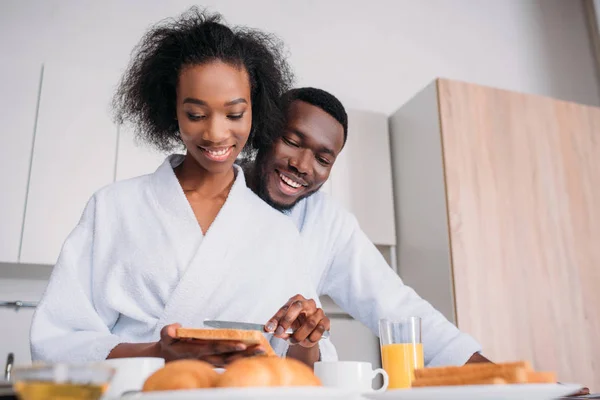 Smiling african american woman spreading butter on toast with boyfriend standing behind
