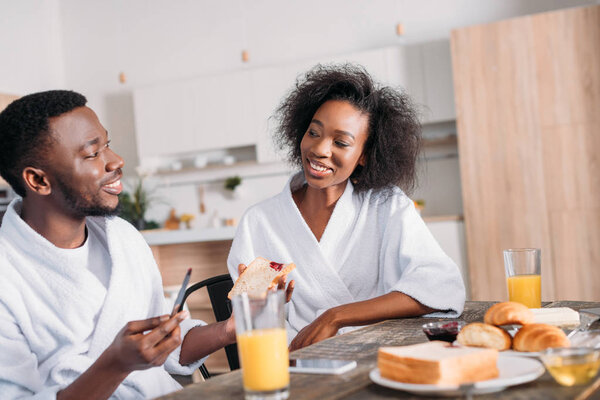 Smiling man spreading jam on toast and girlfriend sitting at table with breakfast 