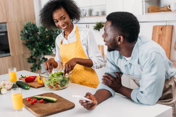 在桌和男朋友站立与智能手机在手的年轻妇女在碗里混合沙拉 — 图库照片