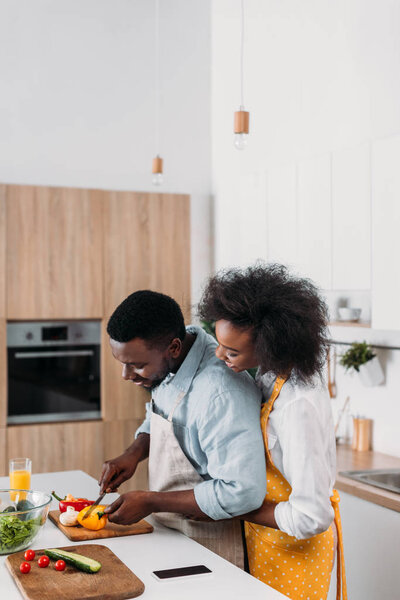 Woman embracing boyfriend while he cutting vegetables on board at table