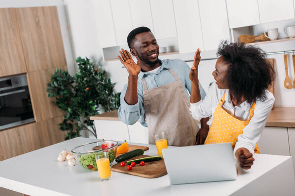 Young couple in aprons giving high five near table with laptop and food