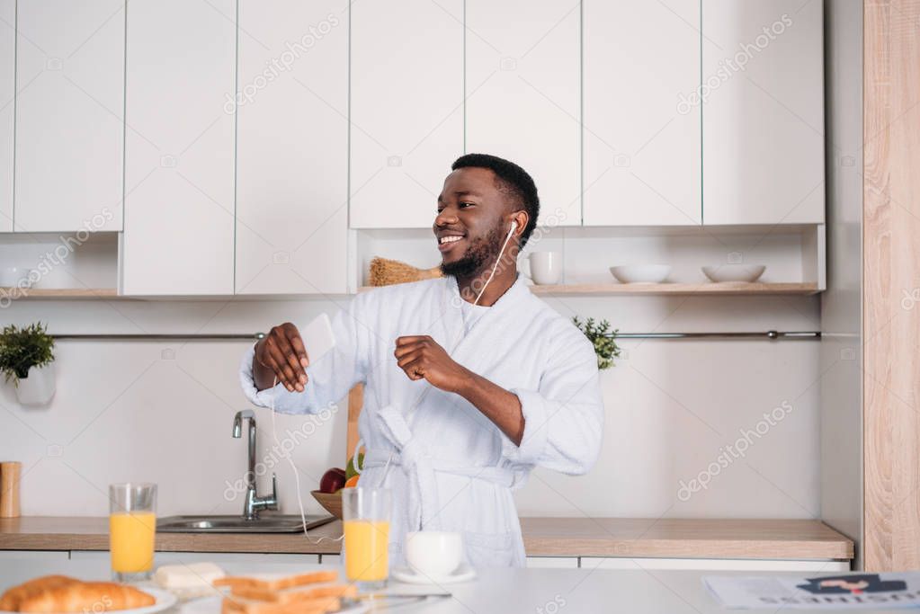 African american man in earphones listening to music from smartphone and dancing in kitchen