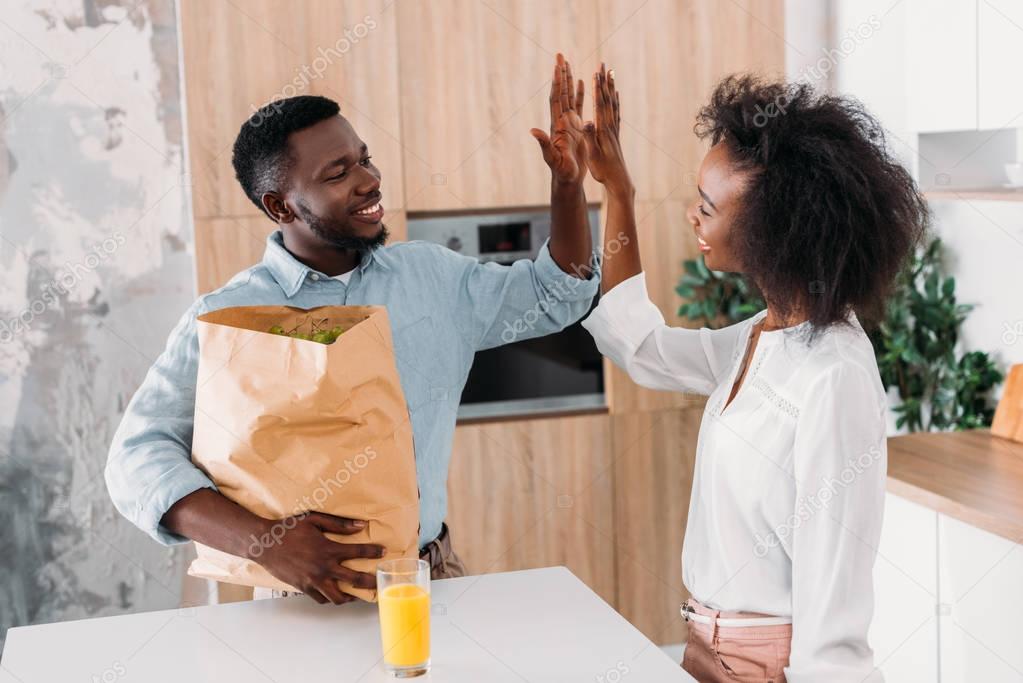 Young man holding paper bag with products and doing high five with girlfriend in kitchen