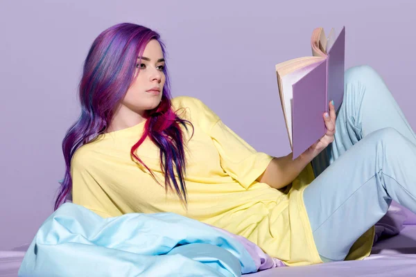 严肃的年轻妇女与五颜六色的头发读书书在床上 — 图库照片