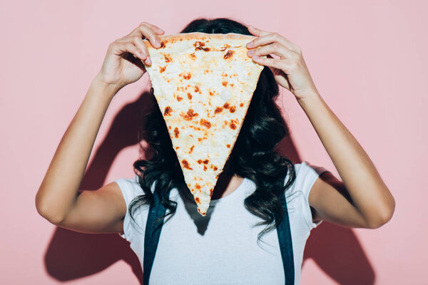 obscured view of woman covering face with piece of pizza on pink background