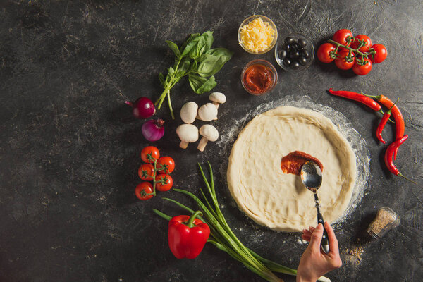 cropped shot of woman putting sauce on raw dough while cooking italian pizza on dark tabletop
