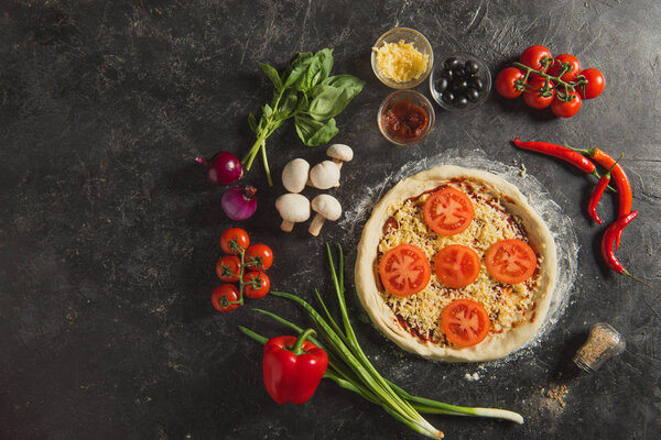 top view of raw italian pizza and fresh ingredients around on dark surface