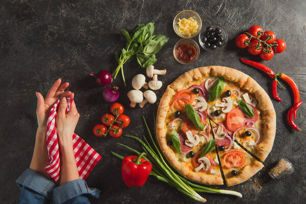 cropped shot of female hands and cooked italian pizza with fresh ingredients on dark tabletop