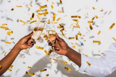 close-up partial view of african american couple clinking glasses of champagne on white with golden confetti clipart