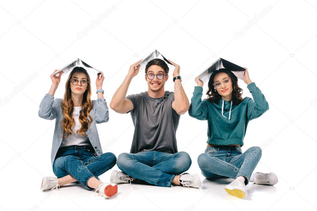 smiling multicultural students sitting with books above heads on white