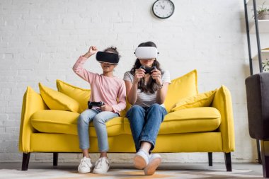 mother and daughter in vr headsets playing video games at home on couch clipart