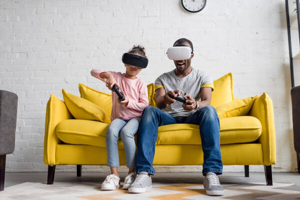 отец и дочь в гарнитурах VR играют дома в видеоигры на диване
