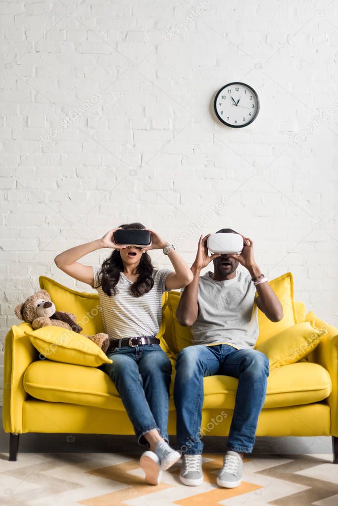 shocked african american couple in virtual reality headsets sitting on couch