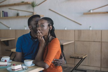 african american man telling secret to shocked girlfriend at table in cafe clipart