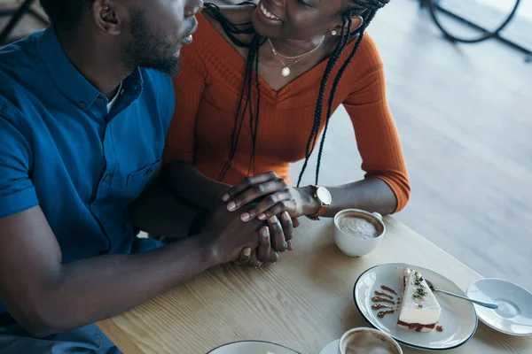 コーヒー ショップでロマンチックな日にテーブルに座って手を繋いでいるアフリカ系アメリカ人のカップルの部分的なビュー  — 無料ストックフォト