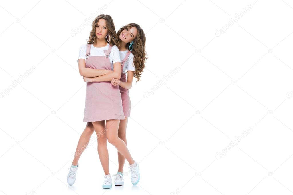 smiling beautiful young twin hugging grimacing sister isolated on white