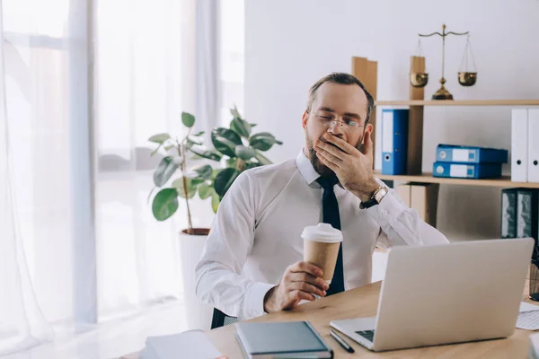 portrait of tired lawyer with coffee to go yawning at workplace in office