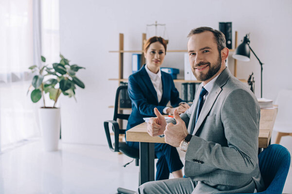 selective focus of smiling businessman showing thumbs up and colleague behind at workplace in office