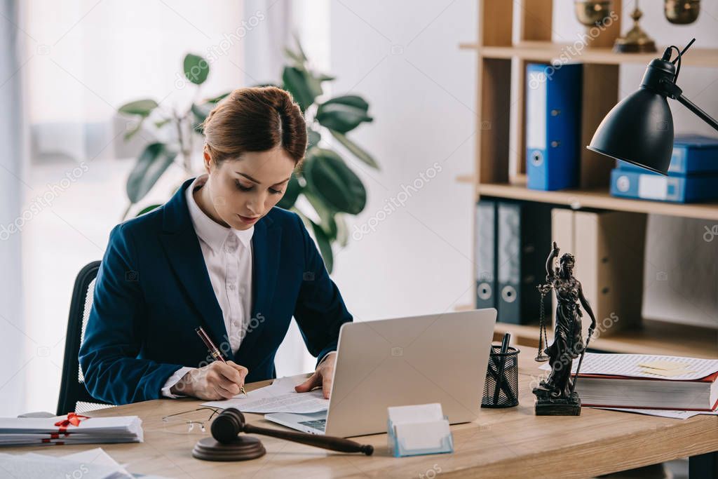 female lawyer in suit at workplace with laptop, gavel and femida in office