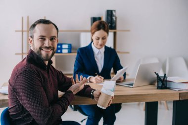 selective focus of smiling client with coffee to go pointing at businesswoman at workplace in office