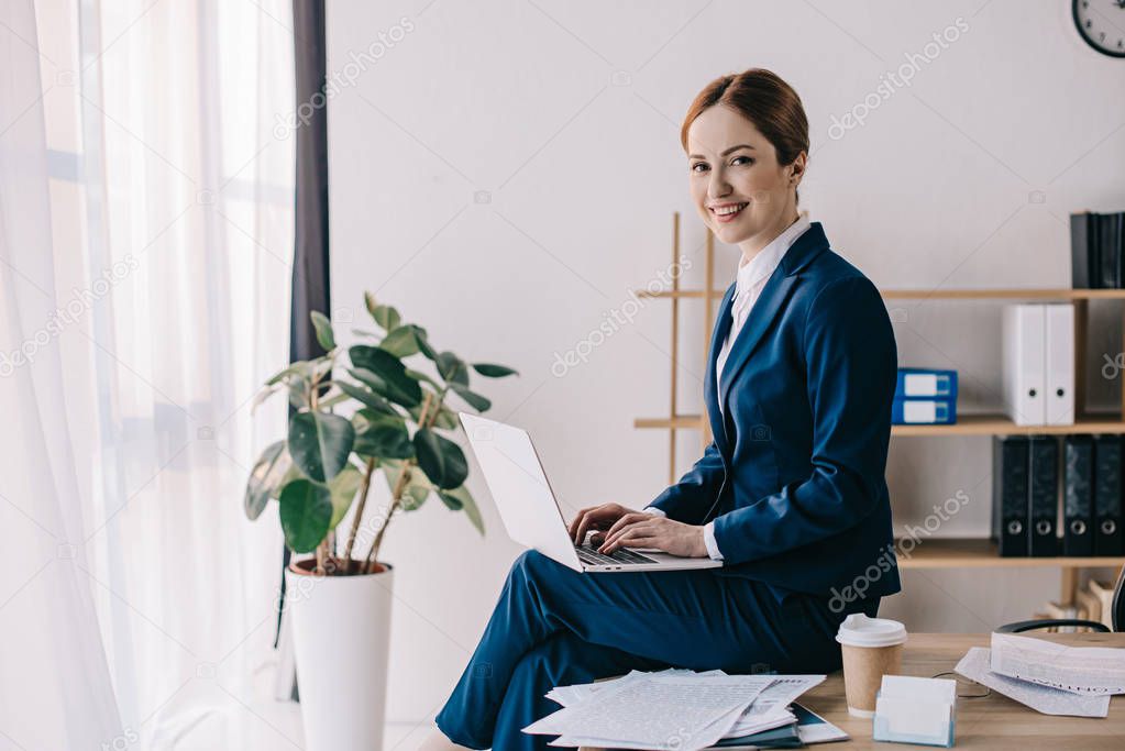 side view of smiling businesswoman with laptop sitting on table in office
