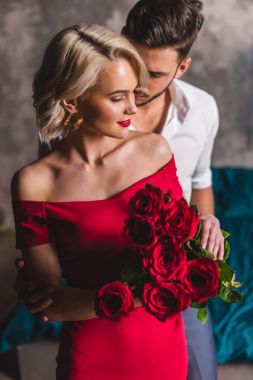 handsome man hugging smiling sensual woman in red dress holding beautiful roses clipart