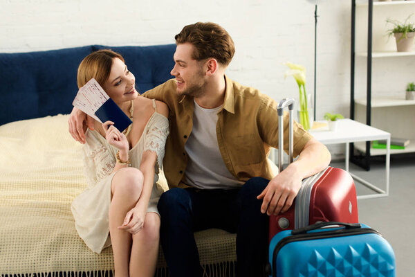 smiling couple with suitcases on bed and holding tickets with passports