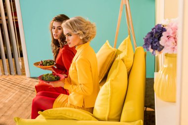 pretty women in retro clothing with vegetables on plates sitting on yellow sofa at colorful room, doll house concept clipart