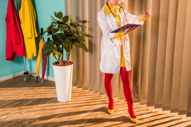 cropped image of retro styled doctor in colorful dress and tights looking at clipboard in clinic clipart