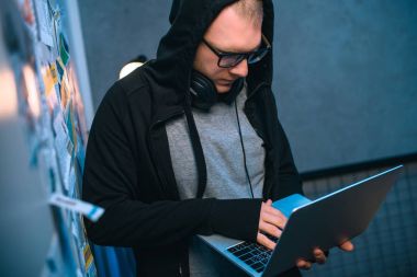 hooded hacker developing malware with laptop in dark room clipart