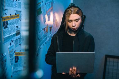 young female hacker with laptop standing in front of wall with newspaper clippings clipart