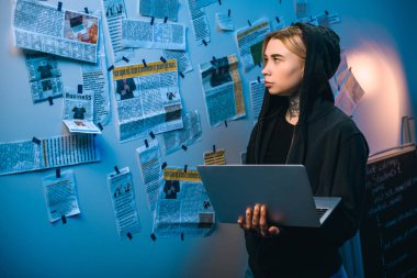 female hacker with laptop standing in front of wall with newspaper clippings clipart