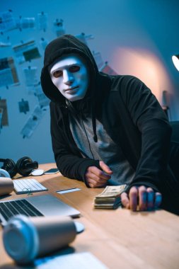 hacker in mask with stack of money on desk clipart