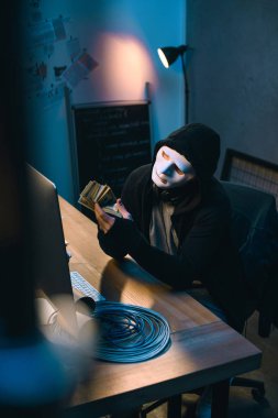 hacker in mask counting stolen money at his workplace clipart