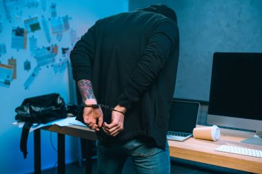 rear view of handcuffed hacker in front of his work desk clipart